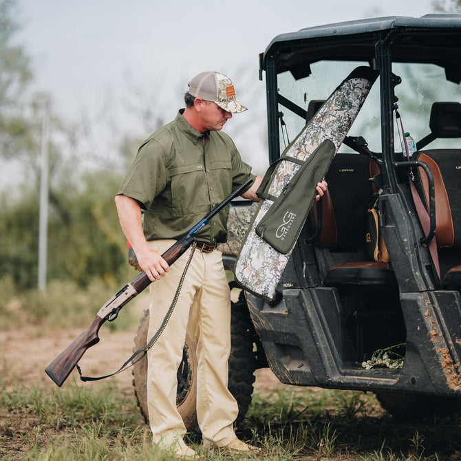 A man in camouflage hunting attire stands next to a black and brown camouflage utility vehicle, holding a gun case in an outdoor field.